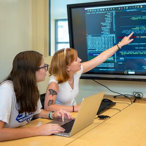 两个学生在一台电脑前学习，电脑上有一个显示数据的大电视屏幕